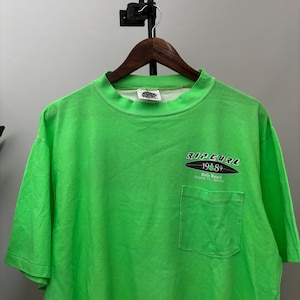 Puede incluir: Camiseta verde brillante con bolsillo y mangas cortas. La camiseta presenta el texto "RIP CURL 1989 Bells Beach Torquay, Vic, Australia" en blanco y negro en la parte delantera. La camiseta est&aacute; colgada en una percha de madera.
