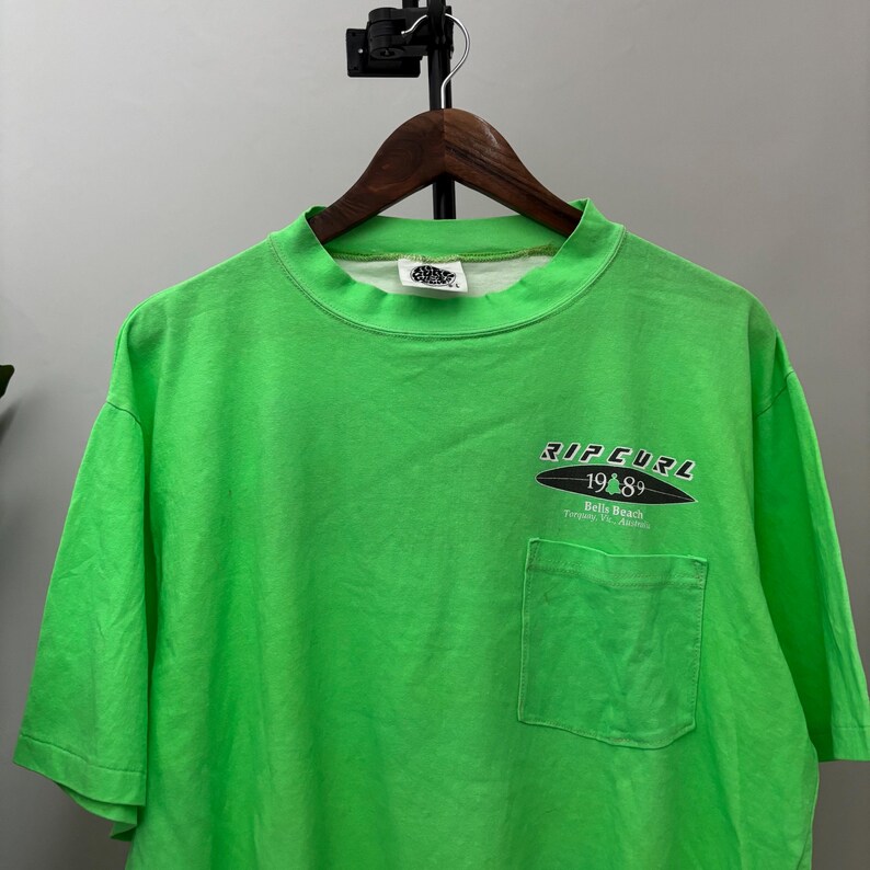 Puede incluir: Camiseta verde brillante con bolsillo y mangas cortas. La camiseta presenta el texto "RIP CURL 1989 Bells Beach Torquay, Vic, Australia" en blanco y negro en la parte delantera. La camiseta est&aacute; colgada en una percha de madera.