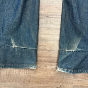May include: Close-up of the bottom of two denim jean legs. The jeans are a faded blue with distressed areas and frayed edges at the hem. The fabric shows signs of wear and tear, giving a vintage look.