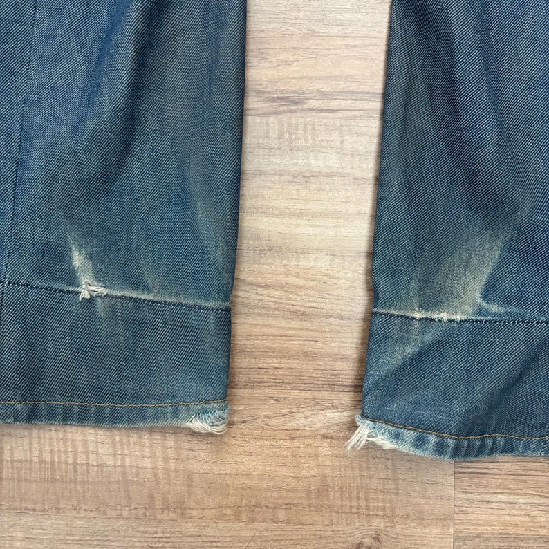 May include: Close-up of the bottom of two denim jean legs. The jeans are a faded blue with distressed areas and frayed edges at the hem. The fabric shows signs of wear and tear, giving a vintage look.
