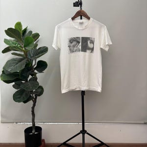 May include: White short-sleeve t-shirt with a black and white graphic print. The graphic features two side-by-side portraits of a person. The t-shirt is displayed on a black stand in front of a gray wall.
