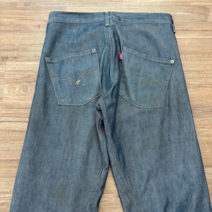 May include: Blue denim jeans with two back pockets and a red tag. The jeans have a classic five-pocket design and a straight leg fit. The jeans are made of a durable denim fabric and are suitable for everyday wear.