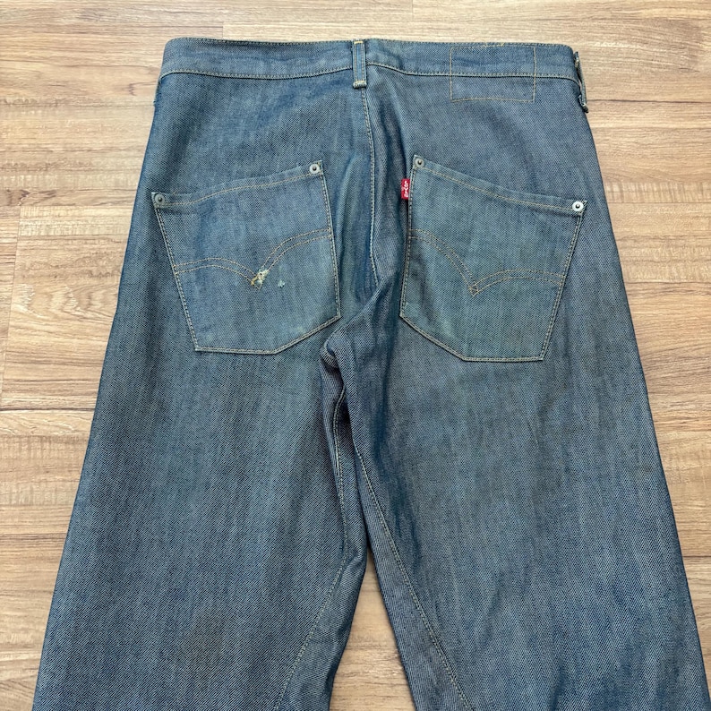 May include: Blue denim jeans with two back pockets and a red tag. The jeans have a classic five-pocket design and a straight leg fit. The jeans are made of a durable denim fabric and are suitable for everyday wear.