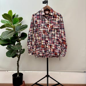 Puede incluir: Camisa de cuadros de manga larga con diseño patchwork, con una mezcla de cuadrados rojos, blancos y azul marino. La camisa, con dos bolsillos en el pecho, se muestra en una percha.