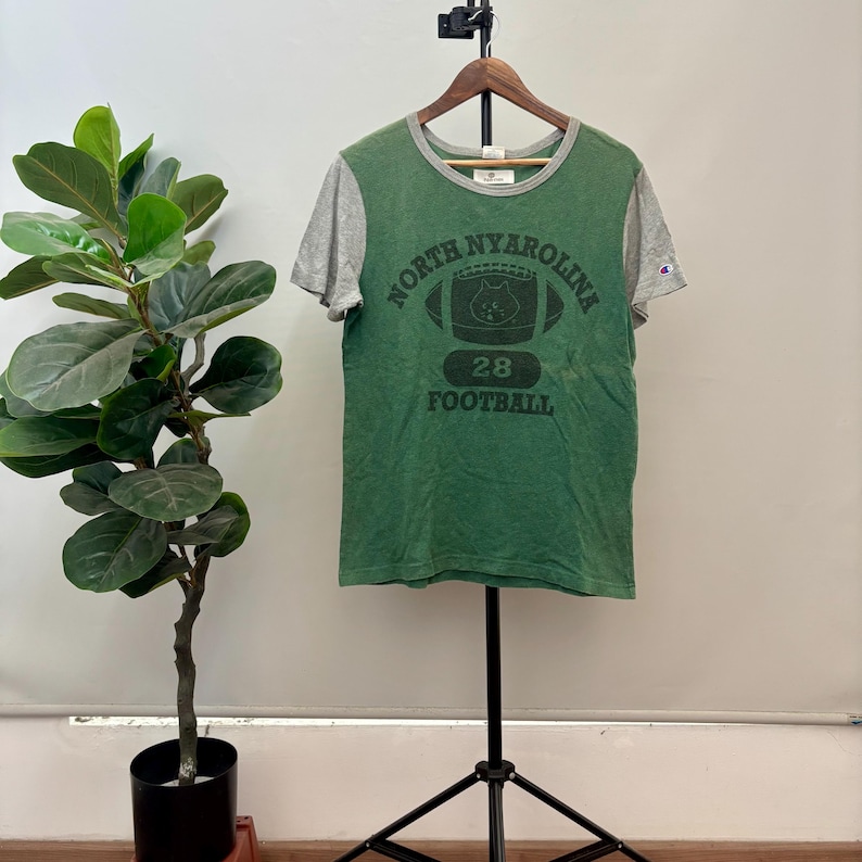 May include: A green and gray short-sleeve t-shirt with a crew neckline. The front features a dark green graphic of a football with a cat face inside, the text NORTH NYAROLINA, the number 28, and the word FOOTBALL. A small Champion logo is on the sleeve.