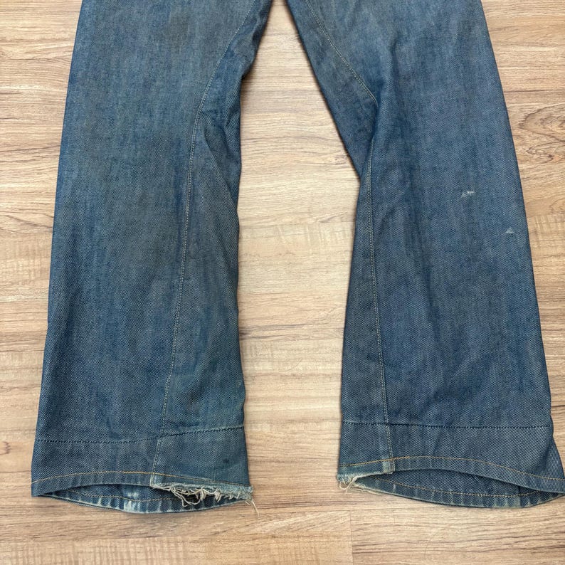 May include: Light blue denim jeans with a wide leg. The jeans feature a classic five-pocket design and a button and zip closure. The hemline is frayed, giving them a vintage appearance.