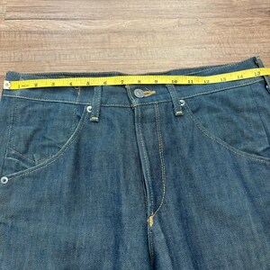 May include: Close-up of blue denim jeans with a button closure and front pockets. A yellow measuring tape shows a 35.5 cm waist measurement. The jeans have visible stitching and a classic design.