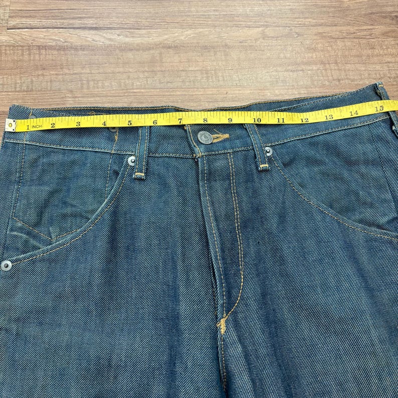 May include: Close-up of blue denim jeans with a button closure and front pockets. A yellow measuring tape shows a 35.5 cm waist measurement. The jeans have visible stitching and a classic design.