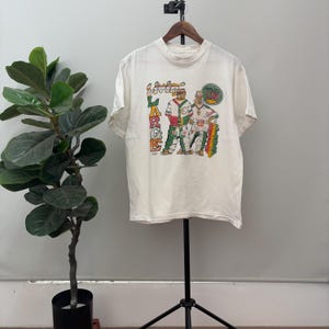 May include: Off-white t-shirt featuring a graphic of two cartoon figures in colorful outfits. The text "Livin' Large" is vertically printed on the left side of the shirt, with the words "Slow Down" in a circle on the right.