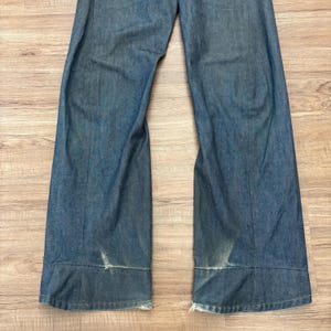 May include: Blue denim trousers with a wide leg cut. The jeans have a faded look and distressed areas near the hem. The fabric appears to be a medium-wash denim.
