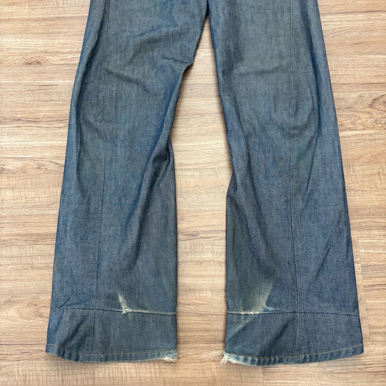 May include: Blue denim trousers with a wide leg cut. The jeans have a faded look and distressed areas near the hem. The fabric appears to be a medium-wash denim.