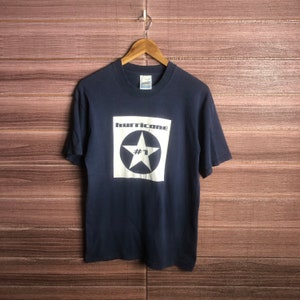 Może przedstawiać: Granatowa koszulka z białym kwadratowym nadrukiem. Grafika zawiera słowo "hurricane" nad białą gwiazdą w czarnym okręgu, z numerem "1" poniżej. Koszulka wisi na drewnianym wieszaku.