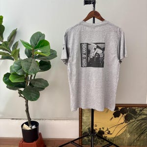 May include: A heather gray t-shirt with a black and white graphic on the back. The graphic features a band performing with the text "SHOW OF HANDS" vertically on the left side. The shirt is hanging on a wooden hanger.