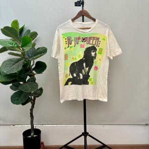 Puede incluir: Camiseta de color crema con estampado gráfico. El estampado presenta el texto "JU-JU QUEEN" en verde y amarillo, con una imagen en blanco y negro de una persona. La camiseta se muestra en una percha.