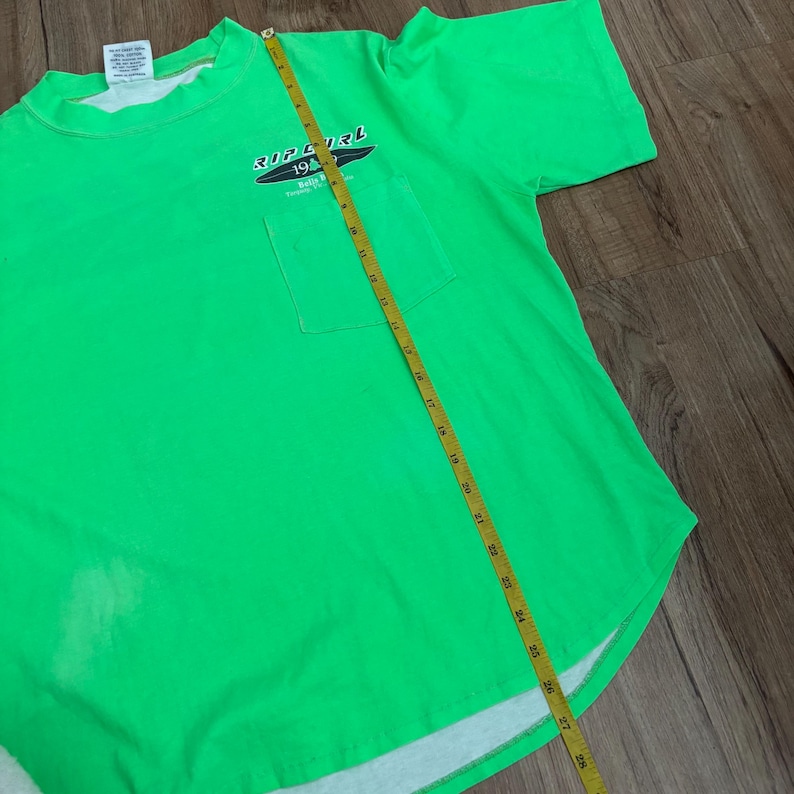Puede incluir: Camiseta verde brillante con bolsillo y el logo de Rip Curl. La camiseta tiene cuello redondo y mangas cortas. Una cinta m&eacute;trica amarilla est&aacute; colocada sobre la camiseta, mostrando una medida de aproximadamente 71 cm.