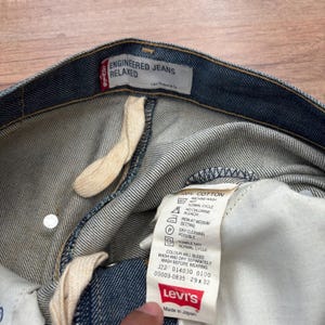 May include: Close-up of blue denim Levi's Engineered Jeans Relaxed. The jeans have a white tag with washing instructions and the Levi's logo. The tag also indicates the size is 29 x 32.