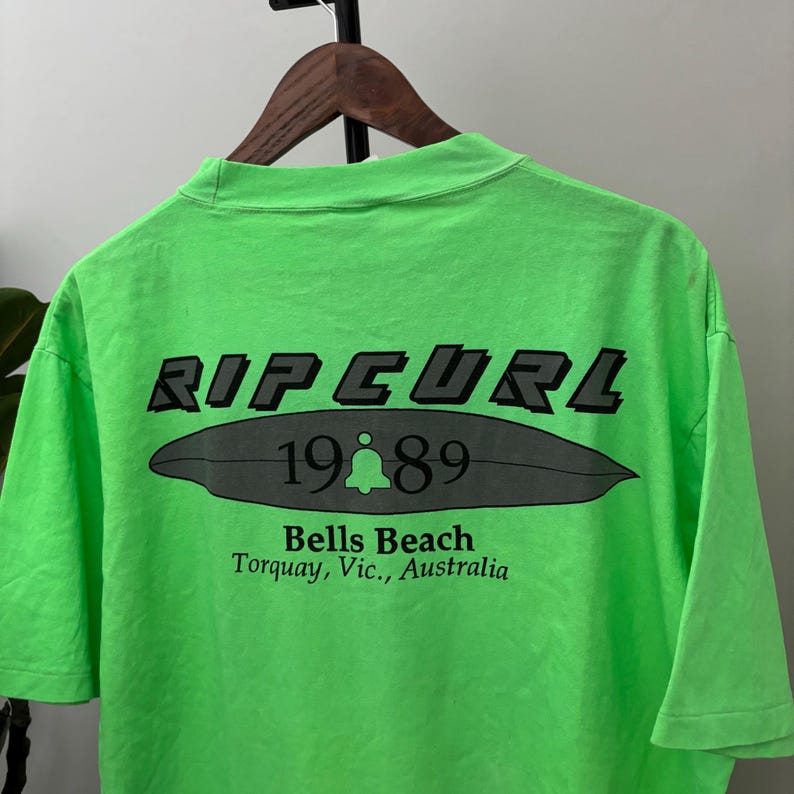 Puede incluir: Camiseta verde brillante con el texto "RIP CURL 1989" sobre un gr&aacute;fico de tabla de surf. Debajo, el texto "Bells Beach Torquay, Vic., Australia". La camiseta est&aacute; colgada en una percha de madera.