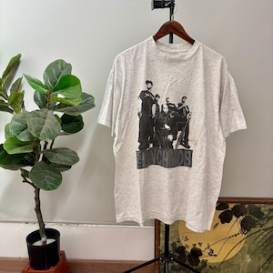 Puede incluir: Una camiseta gris claro con un gráfico en blanco y negro de un grupo de personas. El texto "THE LYNCH MOB" está impreso en un recuadro rectangular en la parte inferior del gráfico. La camiseta está colgada en una percha de madera.
