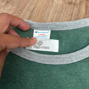 May include: A green t-shirt with a grey crew neckline. The interior collar features a white Champion brand tag labeled size Large and an additional white Ne-net brand tag positioned directly below the Champion label on the garment interior.