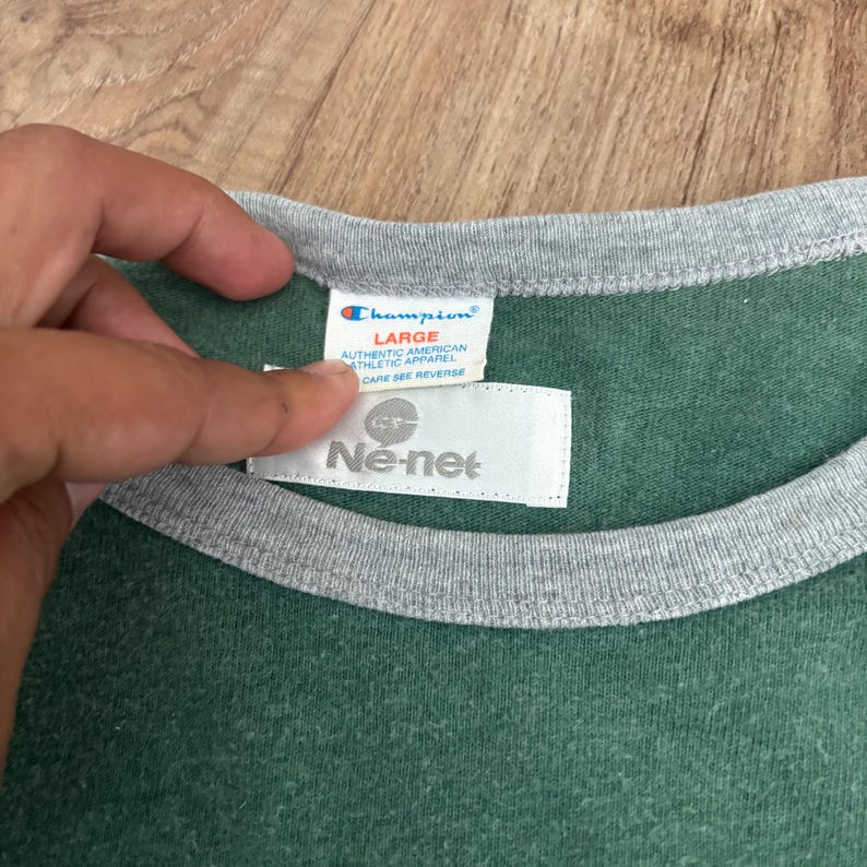 May include: A green t-shirt with a grey crew neckline. The interior collar features a white Champion brand tag labeled size Large and an additional white Ne-net brand tag positioned directly below the Champion label on the garment interior.