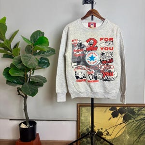May include: A light gray Converse sweatshirt with a graphic design featuring a Converse All Star shoe, text, and cartoon illustrations in red, black, and white. The sweatshirt is hanging on a wooden hanger.