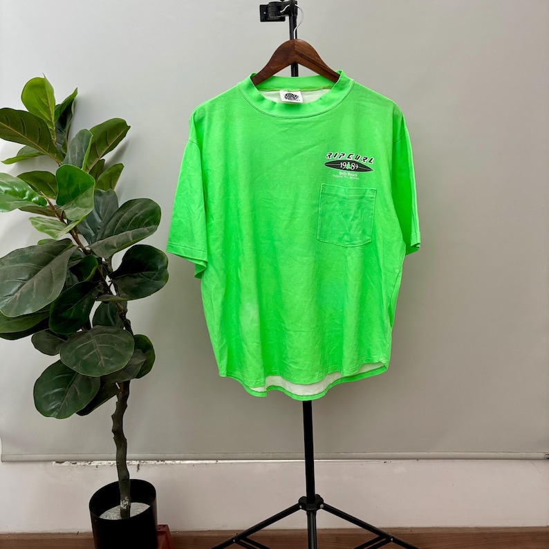 Puede incluir: Camiseta verde ne&oacute;n brillante con un peque&ntilde;o bolsillo y el texto "RIPCURL 1969" en blanco y negro. La camiseta de manga corta tiene cuello redondo y est&aacute; colgada de una percha de madera.