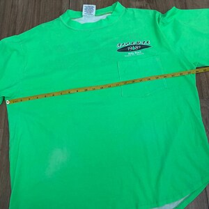 Puede incluir: Camiseta verde brillante con bolsillo y el texto "RIP CURL 1989 Bells Beach". La camiseta se mide con una cinta m&eacute;trica amarilla. La camiseta mide aproximadamente 61 cm de ancho.