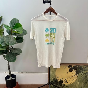 May include: A white short-sleeve t-shirt featuring a graphic print of three small alien characters in blue, green, and yellow, stacked vertically next to the numbers 30, 20, and 10, with the word INVADERS printed in blue at the bottom.