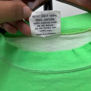 Puede incluir: Primer plano de un cuello de camiseta verde ne&oacute;n con una etiqueta blanca. La etiqueta dice "TO FIT CHEST 100cm, 100% COTTON, WARM MACHINE WASH, DO NOT BLEACH, DO NOT TUMBLE DRY, WARM IRON, MADE IN AUSTRALIA."