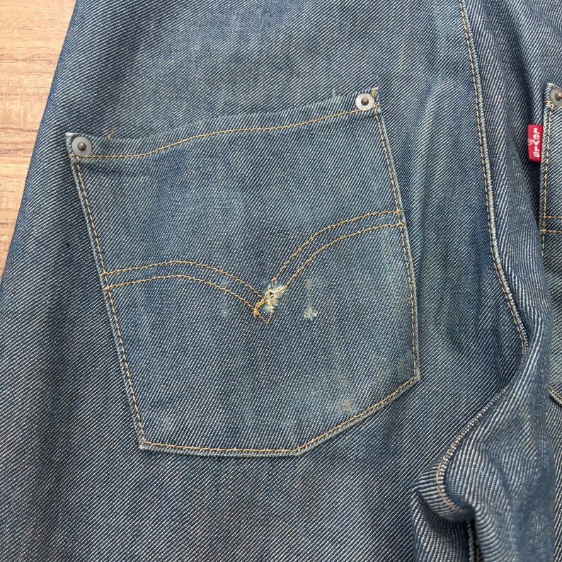 May include: Close-up of blue denim jeans, showing the back pocket with yellow stitching and a small tear. The jeans have a faded look, with a red tag on the side. The denim is a classic, durable material.