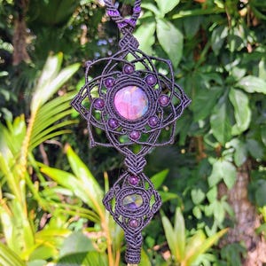 Op de afbeelding: Een handgemaakte macramé ketting met een centrale hanger met een grote, ronde, iriserende steen omringd door kleinere paarse kralen. De ketting heeft een geometrisch ontwerp met een helder kwartskristal aan de onderkant.