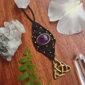 Op de afbeelding: Een handgemaakte macramé ketting met een grote ovale amethiststeen en goudkleurige accenten. De hanger heeft een Keltisch knoopontwerp. De ketting wordt gepresenteerd op een houten oppervlak met kristallen en een varen.