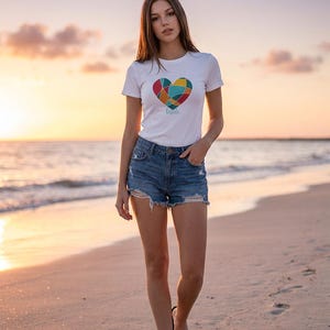 May include: A white t-shirt featuring a colourful heart design and the word "bride". The t-shirt is worn with denim shorts. The person is walking on a sandy beach at sunset, wearing black sandals.