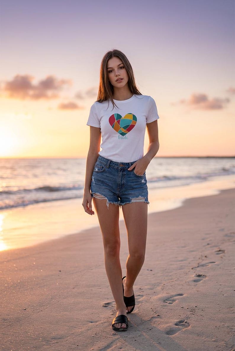 May include: A white t-shirt featuring a colourful heart design and the word "bride". The t-shirt is worn with denim shorts. The person is walking on a sandy beach at sunset, wearing black sandals.