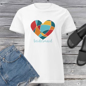 Può includere: T-shirt bianca con un design a cuore geometrico colorato in rosso, arancione, giallo e turchese. La parola "bridesmaid" è scritta in un carattere turchese sotto il cuore. La maglietta è abbinata a pantaloncini di jeans e sandali neri.