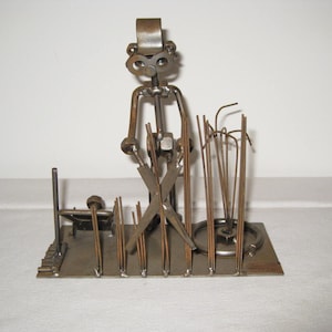 May include: A metal sculpture of a person holding a pair of scissors and standing in front of a row of metal blades, representing grass. The sculpture is made of various metal pieces, including nuts, bolts, and wire.