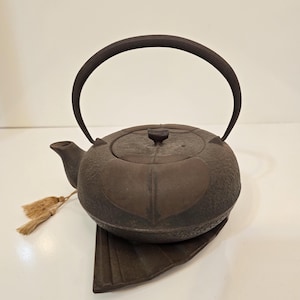 Puede incluir: Una tetera de hierro fundido marrón oscuro con un asa curva y un pequeño pico. La tetera se asienta sobre un salvamanteles a juego en forma de hoja. Una pequeña borla cuelga del lateral. La tetera tiene una superficie texturizada y una tapa con un pequeño pomo.