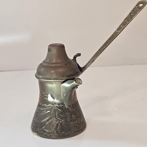Puede incluir: Una pequeña cafetera turca adornada, de color plateado, con un diseño floral y un mango largo. La cafetera tiene una tapa de color cobre.