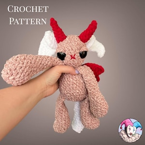 May include: A crocheted plush toy in tan, red, and white, with large black eyes and red horns. The text "CROCHET PATTERN" is visible at the top. The toy has a whimsical, cartoonish design, with a small red cross stitched on its face.