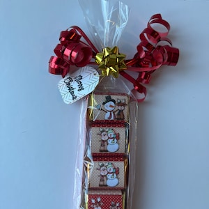 May include: A clear cellophane bag filled with five small wrapped chocolates. The chocolates are decorated with a winter theme featuring snowmen and reindeer. The bag is tied with a red and gold ribbon and a tag that says "Merry Christmas".