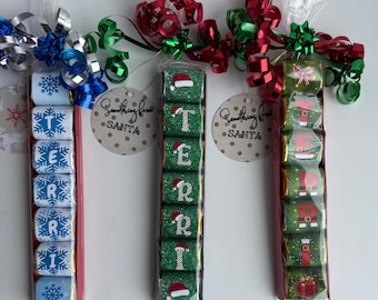 May include: Three gift sets with chocolate candies. One set spells out "TERRI" with snowflake designs, another spells "TER" with Santa hats, and the last spells "TERRI" with elf designs. Each set is decorated with a bow.
