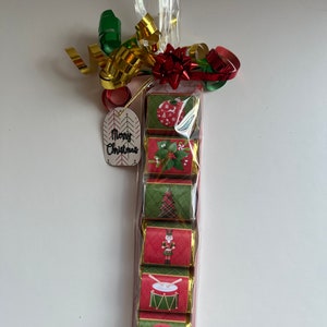 May include: A Christmas-themed gift set of individually wrapped chocolates. The chocolates are in a clear cellophane bag, tied with a red bow, gold and green ribbons, and a tag that says "Merry Christmas". Each chocolate wrapper features a different holiday design.