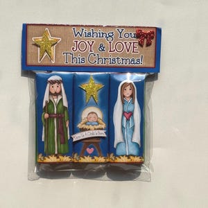 May include: A set of three rectangular chocolate bars in a clear plastic wrapper with a header card that reads Wishing You JOY & LOVE This Christmas! The bars feature illustrations of Joseph, the baby Jesus in a manger, and Mary.