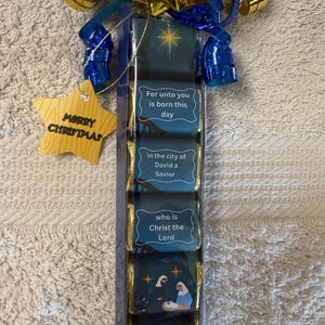 May include: A Christmas gift set featuring a series of chocolate bars with religious imagery and text. The bars are wrapped in gold foil and adorned with blue ribbons and a star-shaped tag that reads "MERRY CHRISTMAS."