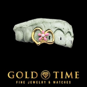 May include: A gold-coloured tooth grill featuring a pink flower design. The grill is set on a grey dental mould against a black background. The text "GOLD TIME FINE JEWELRY & WATCHES" is at the bottom.