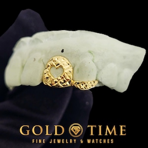 May include: A gold-coloured tooth grill with a heart-shaped cutout and a textured surface. The grill is displayed on a light green dental mould. The image also features the text "GOLD TIME FINE JEWELRY & WATCHES".