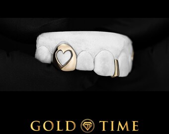 May include: A gold tooth grill piece featuring a heart-shaped cutout design, fitted onto a white dental mold. The grill includes a single heart-accented tooth cover and a smaller gold cap on an adjacent tooth, set against a black background.