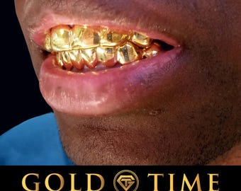 Puede incluir: Un conjunto de parrillas dentales de oro pulido que cubren los dientes superiores e inferiores. Debajo de la boca, una pancarta negra muestra el texto dorado GOLD TIME, un logotipo en forma de diamante con las letras GT y el texto FINE JEWELRY & WATCHES.