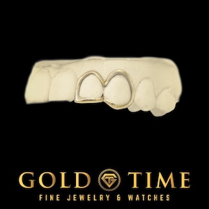 May include: A gold-coloured dental grill, resting on a white dental mold, against a black background. The grill is designed to cover two teeth. The text "GOLD TIME" and "FINE JEWELRY & WATCHES" is at the bottom.