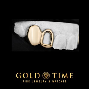 May include: Gold tooth grill with two teeth, one round and one with a hole in the middle. The grill is made of gold and is set on a white mold.